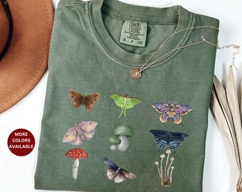 May include: A green t-shirt with a variety of colorful butterflies and mushrooms printed on it. The shirt has the text "COMFORT COLORS" printed on the tag. The text "S - P - CH" is also printed on the tag.