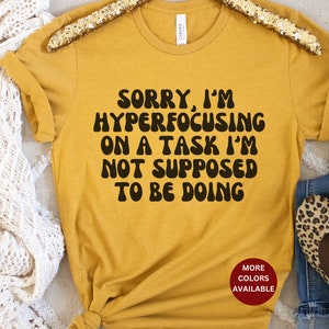 May include: A mustard yellow t-shirt with the text "Sorry, I'm hyperfocusing on a task I'm not supposed to be doing" in black, retro-style lettering. The text is centered on the shirt. The shirt is on a white surface.