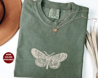 May include: A green t-shirt with a vintage-style moth illustration printed on the front. The shirt is made by Comfort Colors and has a tag that says "S - P - CH".
