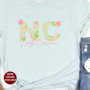 Pu&ograve; includere: Una maglietta blu chiaro con un design floreale che compone la scritta "NC" e le parole "North Carolina" sotto. La maglietta &egrave; su uno sfondo bianco con un paio di sandali, un paio di jeans e una conchiglia.