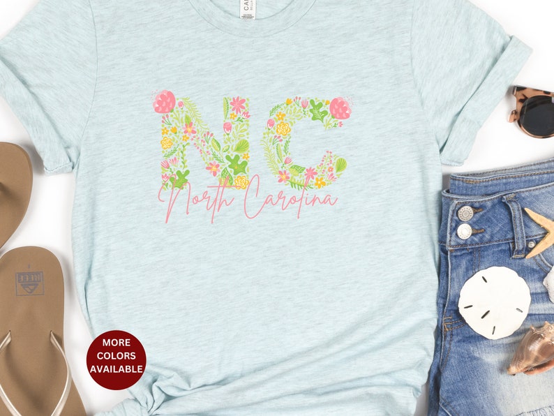 Pu&ograve; includere: Una maglietta blu chiaro con un design floreale che compone la scritta "NC" e le parole "North Carolina" sotto. La maglietta &egrave; su uno sfondo bianco con un paio di sandali, un paio di jeans e una conchiglia.