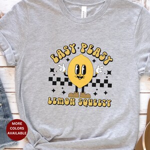 K&ouml;nnte beinhalten: Graues T-Shirt mit einem gelben Zitronen-Cartoon-Charakter, der Schuhe tr&auml;gt und ein Peace-Zeichen macht. Der Text "Easy Peasy Lemon Squeezy" ist auf das T-Shirt gedruckt.