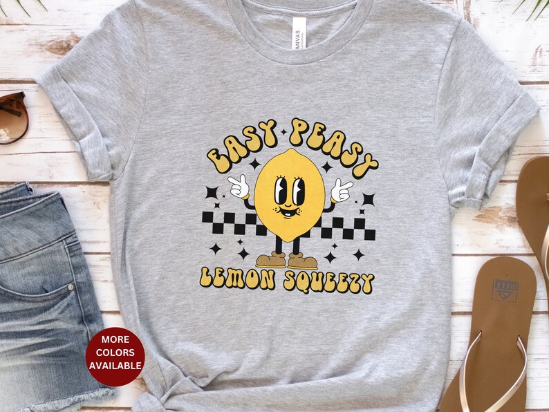 K&ouml;nnte beinhalten: Graues T-Shirt mit einem gelben Zitronen-Cartoon-Charakter, der Schuhe tr&auml;gt und ein Peace-Zeichen macht. Der Text "Easy Peasy Lemon Squeezy" ist auf das T-Shirt gedruckt.