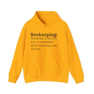 May include: A bright yellow hooded sweatshirt with a front pocket. The text "Beekeeping: Standing in the hot sun in outerwear while holding a can of fire" is printed on the front in black.