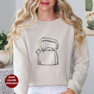May include: A beige sweatshirt with a black and white illustration of a kettle on the front. The text "More Colors Available" is in a red box on the bottom left of the image.