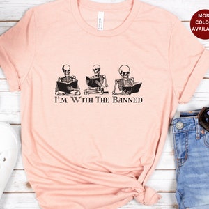 Peut inclure: Un t-shirt rose avec un graphique de trois squelettes lisant des livres. Le texte "I'm With The Banned" est imprim&eacute; sous les squelettes.