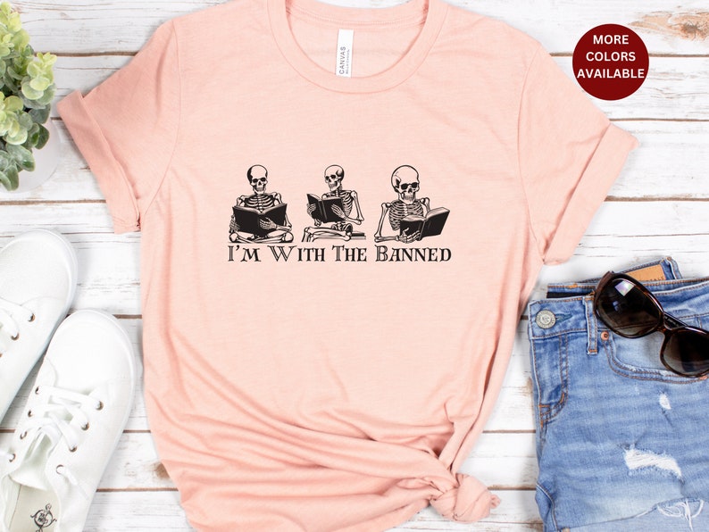 Peut inclure: Un t-shirt rose avec un graphique de trois squelettes lisant des livres. Le texte "I'm With The Banned" est imprim&eacute; sous les squelettes.
