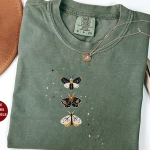 May include: A green t-shirt with a graphic of three moths with gold accents. The shirt has the text "COMFORT COLORS" and "S - P - CH" on the tag.