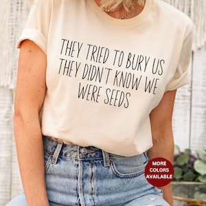 May include: A white t-shirt with the text "They tried to bury us they didn't know we were seeds" printed in black. The shirt is being worn by a person with a light brown complexion and long brown hair. The person is wearing blue jeans.