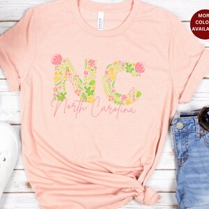 Pu&ograve; includere: Una maglietta rosa con un design floreale che compone la scritta "NC" e le parole "North Carolina" sotto.