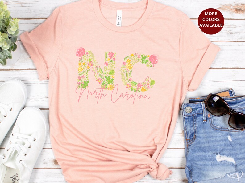 Pu&ograve; includere: Una maglietta rosa con un design floreale che compone la scritta "NC" e le parole "North Carolina" sotto.