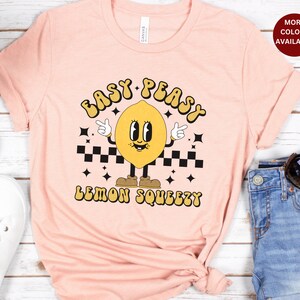 K&ouml;nnte beinhalten: Ein pinkfarbenes T-Shirt mit einem Cartoon-Zitronen-Motiv und dem Text "Easy Peasy Lemon Squeezy".