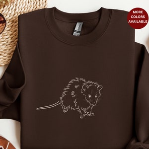 May include: A brown sweatshirt with a white outline of an opossum on the front. The opossum is facing the right side of the image. The sweatshirt is on a white surface.