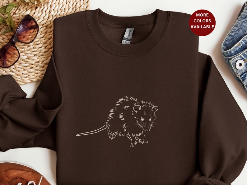 May include: A brown sweatshirt with a white outline of an opossum on the front. The opossum is facing the right side of the image. The sweatshirt is on a white surface.