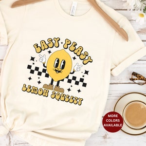 K&ouml;nnte beinhalten: Ein wei&szlig;es T-Shirt mit einer gelben Cartoon-Zitronenfigur, die Schuhe tr&auml;gt und einen Finger hochh&auml;lt. Der Text "Easy Peasy Lemon Squeezy" ist in einer Retro-Schrift auf das Shirt gedruckt.