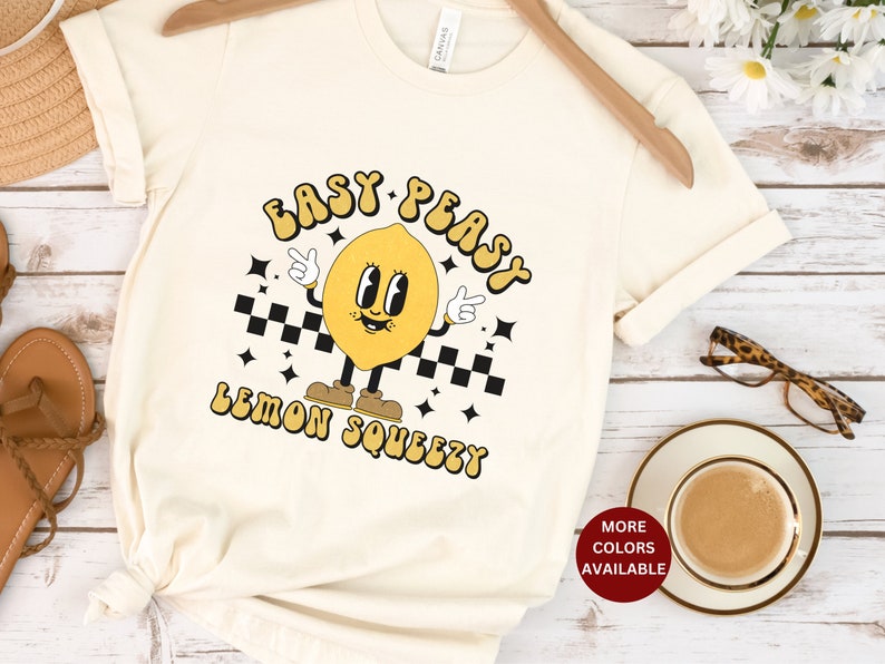 K&ouml;nnte beinhalten: Ein wei&szlig;es T-Shirt mit einer gelben Cartoon-Zitronenfigur, die Schuhe tr&auml;gt und einen Finger hochh&auml;lt. Der Text "Easy Peasy Lemon Squeezy" ist in einer Retro-Schrift auf das Shirt gedruckt.