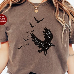 May include: A brown t-shirt with a black silhouette of a raven with constellations on its wings. The raven is flying with its wings spread and there are smaller silhouettes of birds flying behind it.