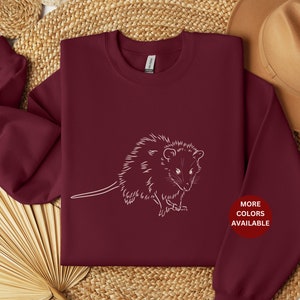 May include: Burgundy sweatshirt with a white outline of a possum. The text "MORE COLORS AVAILABLE" is in a white circle on the bottom right of the sweatshirt.
