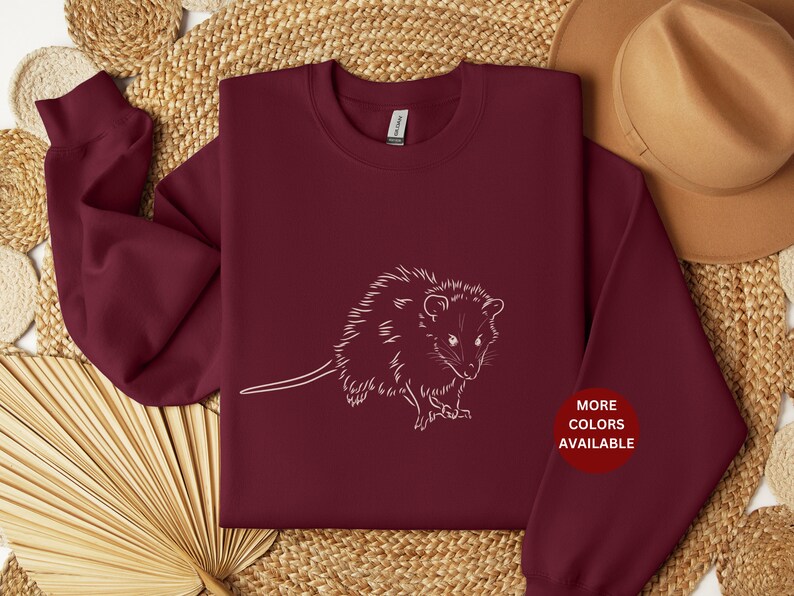 May include: Burgundy sweatshirt with a white outline of a possum. The text "MORE COLORS AVAILABLE" is in a white circle on the bottom right of the sweatshirt.