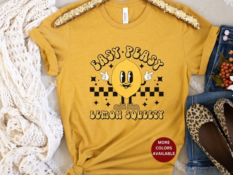K&ouml;nnte beinhalten: Ein senfgelbes T-Shirt mit einer Retro-Grafik eines l&auml;chelnden Zitronen mit dem Text "Easy Peasy Lemon Squeezy".