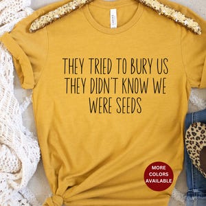 May include: A mustard yellow t-shirt with the text "They tried to bury us they didn't know we were seeds" printed in black. The text is centered on the shirt. The shirt is on a white surface. The text "More Colors Available" is printed in red on a white circle.