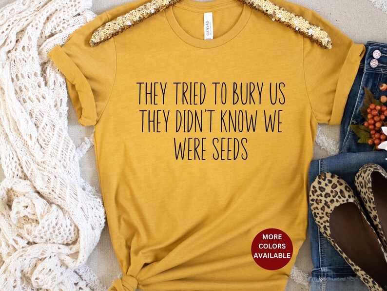 May include: A mustard yellow t-shirt with the text "They tried to bury us they didn't know we were seeds" printed in black. The text is centered on the shirt. The shirt is on a white surface. The text "More Colors Available" is printed in red on a white circle.