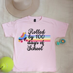 May include: Pink t-shirt with a rainbow design and the text "Rolled by 100 days of school". A cartoon roller skate is on the left side of the design.