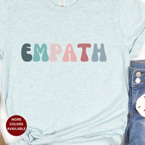 May include: A light blue t-shirt with the word "EMPATH" printed in colorful, retro-style letters. The letters are in shades of teal, green, pink, red, and blue.