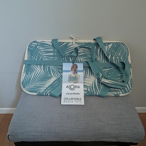 May include: A teal and white collapsible beach tote with a palm leaf pattern. The tote has teal straps and a tag that reads "Aloha" and "Collapsible Beach Tote."