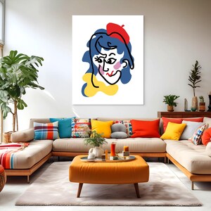 Puede incluir: Una impresión artística enmarcada que muestra un retrato abstracto y colorido de una persona con cabello azul, boina roja y bufanda amarilla. La obra de arte se exhibe en una sala de estar moderna con un sofá seccional beige y cojines coloridos.