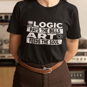 Peut inclure: T-shirt noir avec l'inscription blanche "LOGIC PAYS THE BILLS ART FEEDS THE SOUL". Le t-shirt présente une icône d'ordinateur, une palette de peinture et un pinceau. La personne porte un pantalon marron et une ceinture marron.
