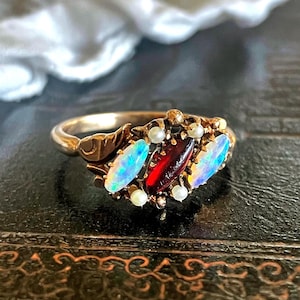 Könnte beinhalten: Ein goldener Ring mit drei ovalen Edelsteinen: zwei Opalen mit schillernden Blau- und Grüntönen und einem tiefroten zentralen Edelstein. Kleine Perlen und goldene Akzente umgeben die Edelsteine. Der Ring hat ein detailliertes, verziertes Design.