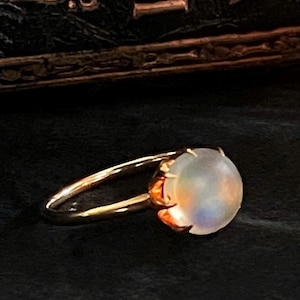 Könnte beinhalten: Ein Goldring mit einem runden, im Cabochon-Schliff geschliffenen Mondstein in einer Krappenfassung. Der Edelstein hat ein durchscheinendes, milchig-weißes Aussehen mit dezenten blauen und orangefarbenen irisierenden Reflexen.