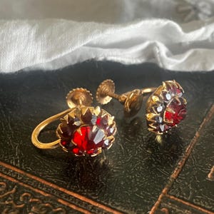 May include: A pair of vintage gold-tone screw-back earrings. Each earring features a cluster of faceted, deep red gemstones, surrounded by smaller, lighter-coloured stones. The earrings are displayed on a dark surface with a white cloth in the background.