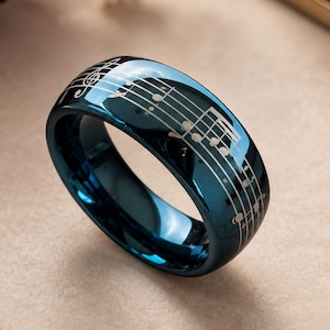 May include: A polished dark blue ring featuring a laser-engraved musical staff with notes and a treble clef wrapping around the outer band. The ring has a smooth, reflective surface that catches the light, showcasing the intricate white musical notation.