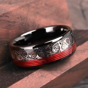 May include: A black ring with a silver skull and swirl pattern and a red stripe. The ring is made of a shiny, dark material and is set against a wooden surface. The ring is a piece of jewelry.
