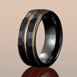 Op de afbeelding: Zwarte ring met een getextureerd, geometrisch patroon en een goudkleurige streep. De ring heeft een gepolijste, glanzende afwerking en een goudkleurige binnenband. Het is een sieraad.