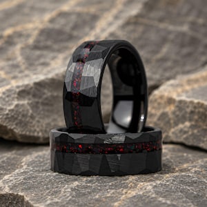May include: Two black rings with a hammered, faceted texture and a central inlay of crushed red and dark iridescent material. The rings feature a dark, matte finish and a distinct geometric pattern across the outer surface of the bands.