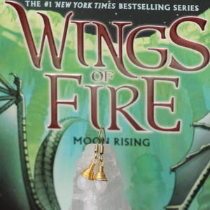 May include: A pair of gold-colored earrings with a wire-wrapped design and a white, round stone. The earrings are displayed in front of a book titled "Wings of Fire: Moon Rising" by Tui T. Sutherland.