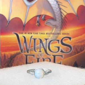 May include: A silver-toned wire-wrapped ring featuring a single round, iridescent white stone at its center. The band is constructed from thin, coiled metal wire, creating a textured appearance around the central gemstone setting.