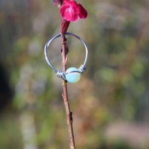 May include: A silver-colored wire ring featuring a single, round, iridescent opal-like bead at the center. The metal wire is wrapped around the bead and twisted to form the band, creating a simple, circular design that rests on a plant stem.