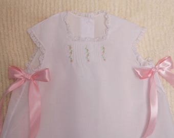 Könnte beinhalten: Ein weißes ärmelloses Kleid mit einem quadratischen Ausschnitt mit Spitzenbesatz, zarten vertikalen Falten und kleiner Blumenstickerei. Die Seiten sind mit großen, hellrosa Satinbändern akzentuiert, die an der Taille zu Schleifen gebunden sind.