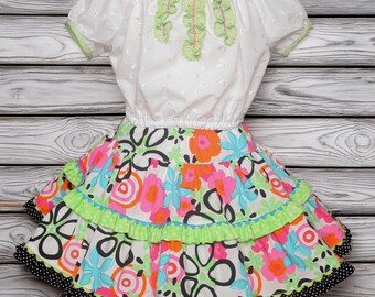 Könnte beinhalten: Ein zweiteiliges Outfit bestehend aus einer weißen kurzärmeligen Bluse aus Lochstickerei mit grüner Verzierung und vertikalen Rüschen, kombiniert mit einem Stufenrock mit buntem Blumenmuster in Pink, Orange, Blau und Schwarz mit Punkten.