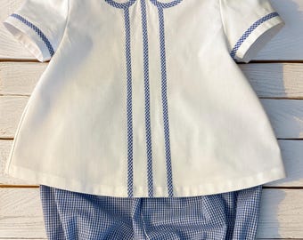 Könnte beinhalten: Ein weißes Kurzarmhemd mit rundem Kragen und drei vertikalen blau-weißen Vichy-Karostreifen auf der Vorderseite, kombiniert mit einer passenden blau-weißen Vichy-Karo-Bloomer-Shorts mit elastischen Beinabschlüssen.