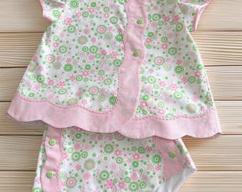 Könnte beinhalten: Ein zweiteiliges Set bestehend aus einem kurzärmeligen Oberteil und einer passenden Hose. Beide Teile bestehen aus weißem Stoff mit einem rosa-grünen Blumenmuster, akzentuiert durch einen rosa Wellensaum und kleine hellgrüne Knöpfe.
