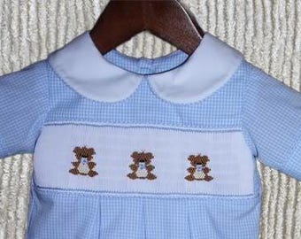Könnte beinhalten: Ein hellblau-weiß kariertes Kurzarmhemd mit einem einfarbigen weißen Rundkragen und einem zentralen weißen gesmokten Einsatz. Der Einsatz ist mit drei gestickten braunen Teddybären verziert, die jeweils eine kleine blaue Fliege tragen.