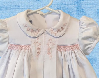 Könnte beinhalten: Ein weißes Kleid mit kurzen Ärmeln, das einen abgerundeten Kragen mit zarter rosa Blumenstickerei, ein zentrales vertikales Paneel mit passenden rosa Blumenmotiven und horizontale Reihen rosa Smok-Stickerei auf der Brust aufweist.