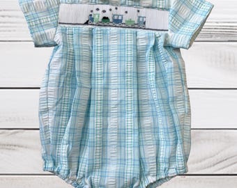 Könnte beinhalten: Ein hellblau-weißer karierter Strampler mit weißem Bubikragen und kurzen Ärmeln. Das Brustteil zeigt ein handgesticktes Smock-Muster mit einer Reihe kleiner, in Blau- und Grüntönen gestickter Züge.