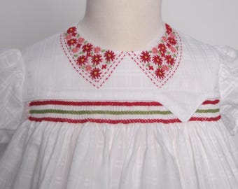 Könnte beinhalten: Ein weißes Kleid mit dezentem Karomuster, kurzen Puffärmeln und einem mit roten und rosa Blumen bestickten Bubikragen. Das Oberteil ist mit horizontalen Smok-Details in abwechselnd roten und grünen Garnfarben versehen.