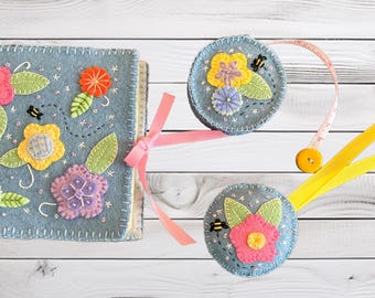 Könnte beinhalten: Ein hellblaues Filzbuch mit gestickten Blumen, grünen Blättern und kleinen Bienen, gebunden mit einem rosa Band. Daneben liegen zwei runde Filzornamente mit passenden Blumenmotiven und ein gelbes Maßband.